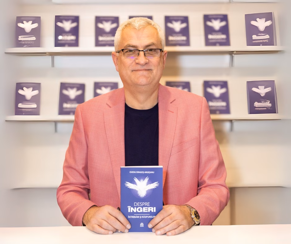 Dragoș Ovidiu Argeșanu, fotografiat în interior. Ține în mâini cartea „Despre îngeri. Întrebări și răspunsuri”, în fața unui raft pe care sunt expuse mai multe exemplare ale aceluiași volum.