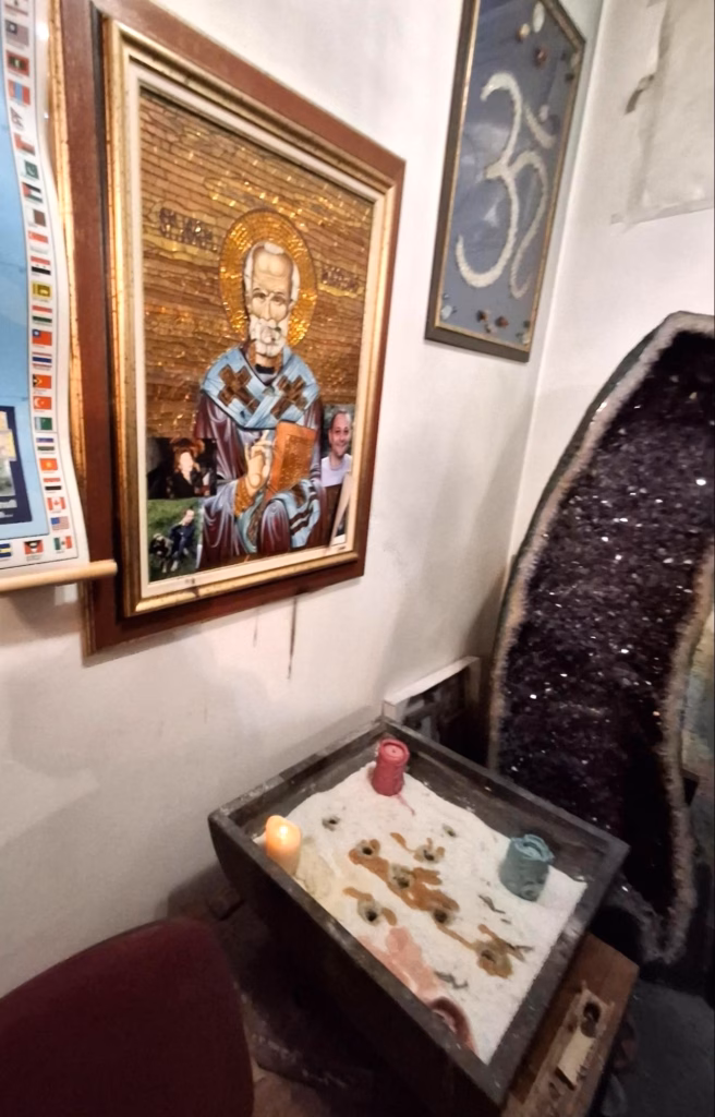 Colț de cameră din cabinetul lui Argesanu: icoană înrămată modificată cu fotografii inserate, simbol „Om” pe perete, o geodă mare de cristal și un suport cu nisip, lumânări și urme de ceară, sugerând un spațiu de practici ezoterice.