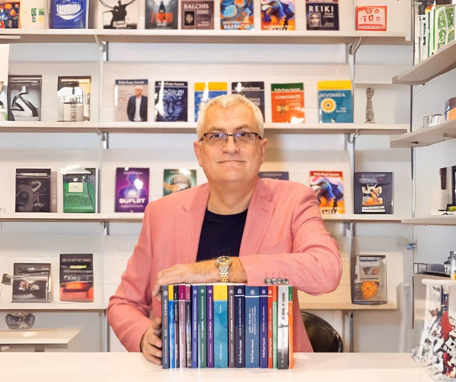 Ovidiu Dragoș Argeșanu fotografiat într-un spațiu cu rafturi pline de cărți, sprijinindu-se pe un teanc de volume proprii expuse pe masă.