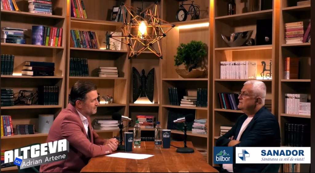Cadru dintr-un interviu în studio: Ovidiu Dragoș Argeșanu stă la masă, față în față cu moderatorul Adrian Artene, într-un decor cu bibliotecă; pe ecran apar logo-urile „Altceva” și „Sanador”.
